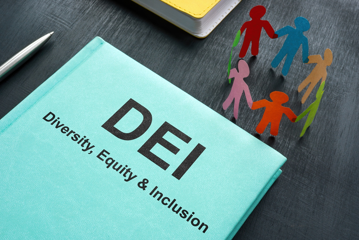 A book titled "DEI: Diversity, Equity, and Inclusion sits on a desk near a ring of multicolored paper dolls standing as if holding hands in a circle. Nearby a pen, other folders/books